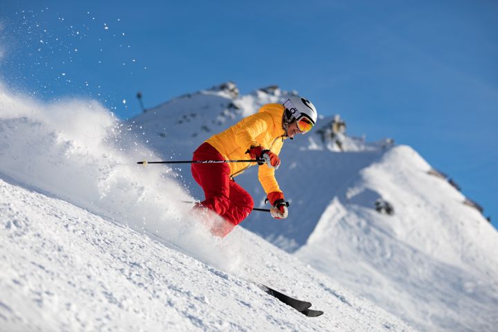 En person står på ski ned en snødekket bakke med blå himmel i bakgrunnen. Personen har på seg en gul jakke og røde bukser.