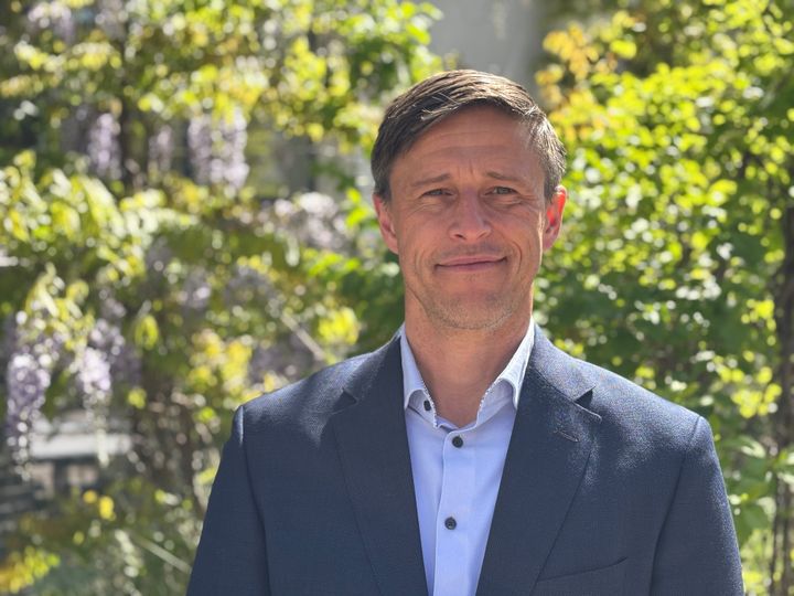 En mand i jakkesæt står foran grønne træer.