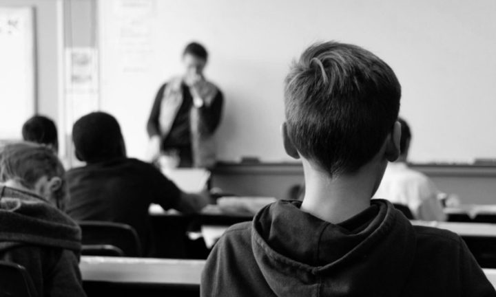 Foto viser ryg af skoleelever i et klasseværelse, hvor læreren står foran tavlen