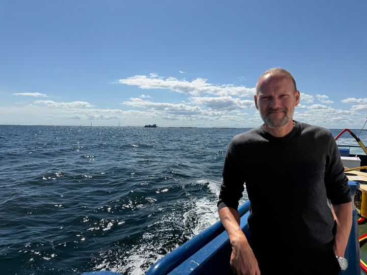 René Christensen, direktør for Dansk Akvakultur Producentorganisation, står på en båd med havudsigt under en solrig himmel.