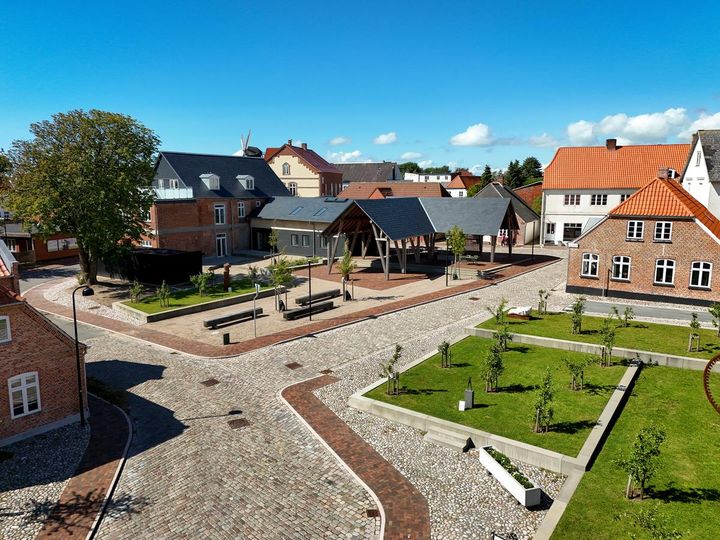 Billede af en brostensbelagt byplads med moderne lamper, træer og røde murstenshuse i baggrunden under en blå himmel.