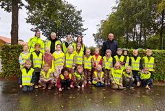 Gruppefoto af børn og voksne iført sikkerhedsveste foran grøn hæk.