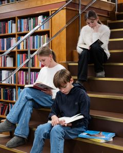 Tre børn sidder og læser bøger på en trappe i et bibliotek.