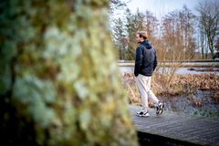 En mand går langs en træbro i en naturskøn omgivelser med træer og vand i baggrunden.