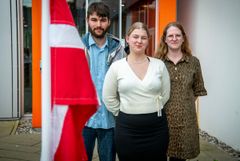 En gruppe på tre unge mennesker står foran en bygning. En rød og hvid flag er i forgrunden.