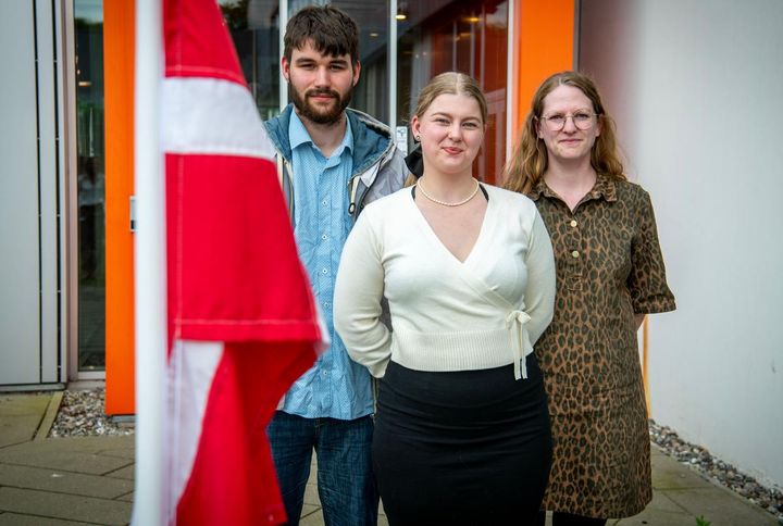 En gruppe på tre unge mennesker står foran en bygning. En rød og hvid flag er i forgrunden.