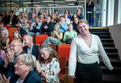 En kvinde går smilende op ad en trappe i en sal fyldt med klappende tilskuere.