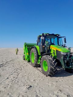 En grøn traktor med tilknyttet rengøringsmaskine kører på en strand under en klar blå himmel.