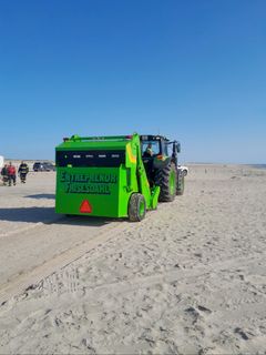 Et grønt rengøringskøretøj arbejder på en sandstrand under en blå himmel.