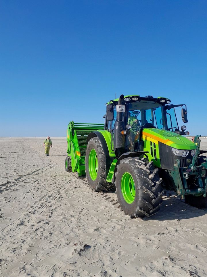 En grøn traktor med en rengøringsmaskine fjerner olieperler fra stranden på Rømø. En person går i baggrunden.