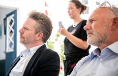 Billede af tre personer, hvor to mænd sidder og lytter opmærksomt, mens en kvinde står med en telefon i hånden.