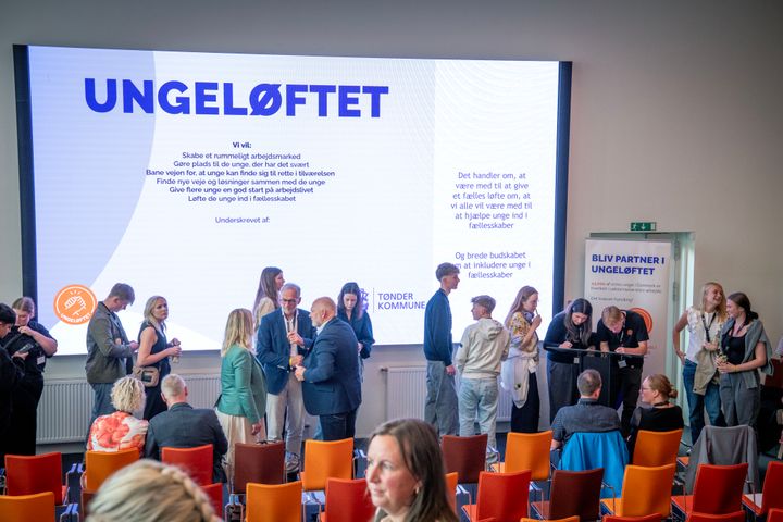 Gruppe af mennesker står i et lokale og underskriver et stort banner med teksten "UNGELØFTET".