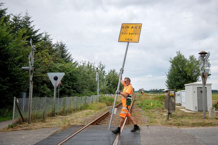 Mand i orange arbejdstøj opsætter gult advarselsskilt ved jernbaneoverskæring.