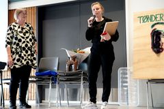 Kvindelig modtager taler med mikrofon og holder et diplom på en scene, mens en anden kvinde kigger på.