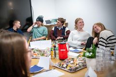 Fem personer sidder ved et bord med mad og drikke. De smiler og ser ud til at have en afslappet samtale i et mødelokale.