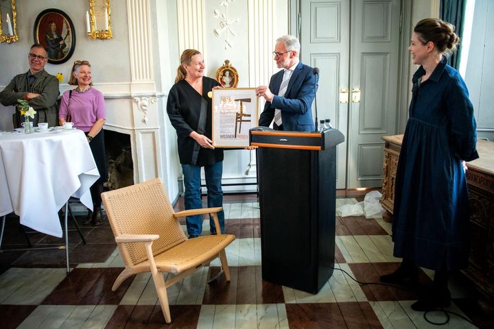 Person modtager en pris ved en ceremoni i et elegant rum. En Wegner-stol er i forgrunden.