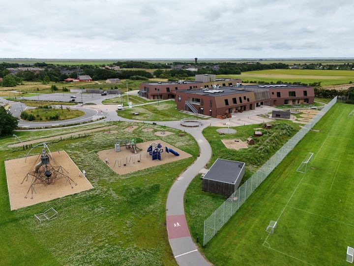 Luftfoto af en moderne skolebygning med legeplads og sportsbaner omgivet af marker.