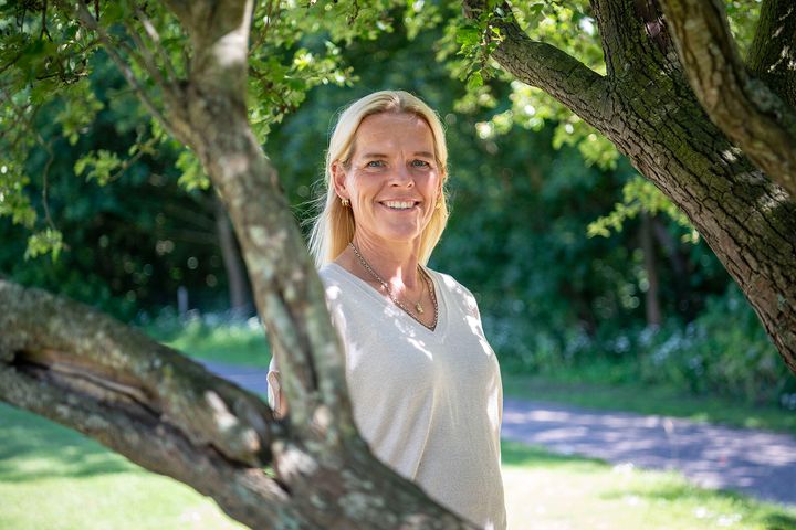 En kvinde står udendørs mellem træer og smiler til kameraet.