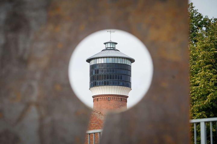 Et rundt udkigshul i en rusten metalplade med udsigt til et rundt tårn med glasvinduer og en kuppel på toppen, omgivet af grønne træer.