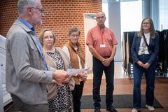 Fem personer står sammen indendørs, mens en person taler og holder nogle papirer.