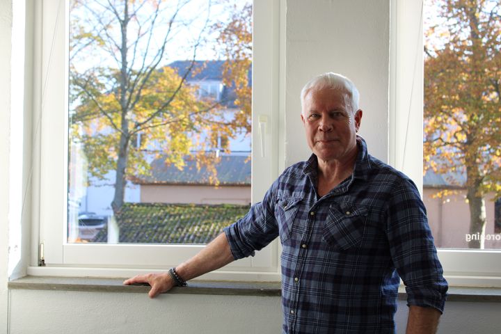 En mand står ved et vindue med træer udenfor. Han har kort, gråt hår og en ternet skjorte.