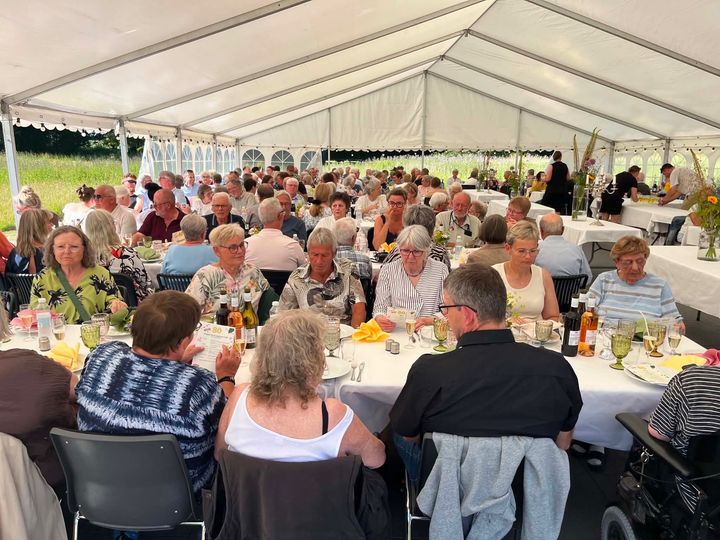 Beboere sidder ved lange borde under et stort festtelt og nyder en sankthansfest.