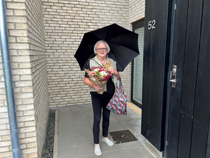 En kvinde står foran sin nye bolig med en paraply og en buket blomster.