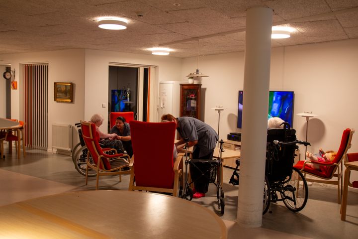 En dagligstue med røde lænestole og en enkelt ældre borger i kørestole. Alle stole er vendt mod fjernsynet på væggen, og en medarbejder i blå dragt bøjer sig ind over en af stolene.