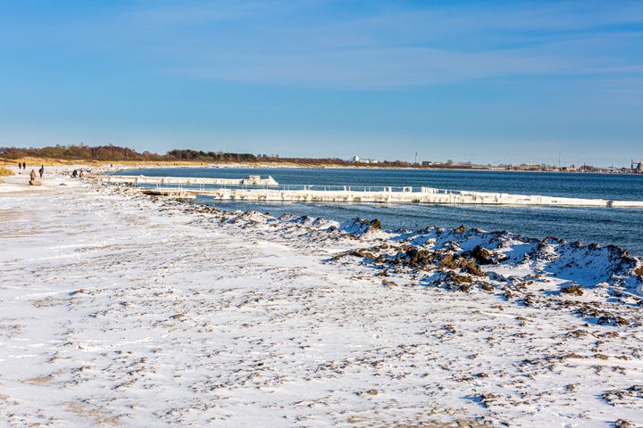 En snedækket strandkant der møder havet. En badebro går ud fra standen og ud. Bagerst kan man i horisonten ane Advedøre Værket.