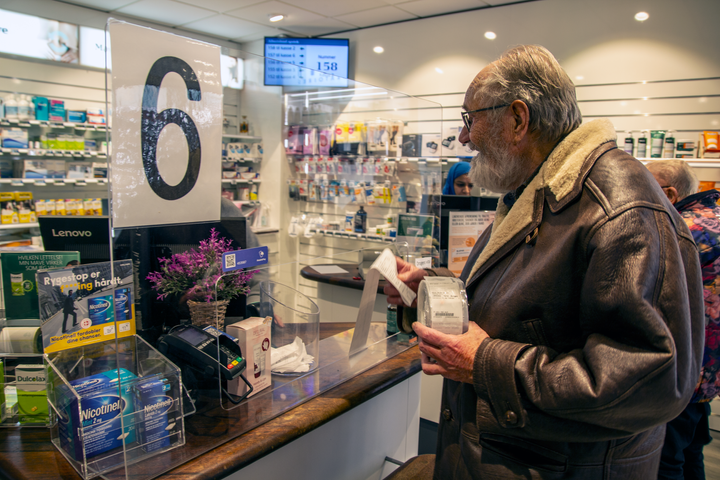 En ældre mand i oilskinsjakke står ved en disk på et apotek med sit køb i hånden.