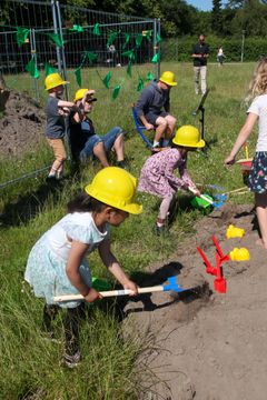 Børn iført gule byggepladshjelme graver i jorden med skovle, mens voksne kigger på.