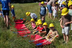 Børn med gule byggehatte sidder i rækker af røde trillebørre og leger udendørs.