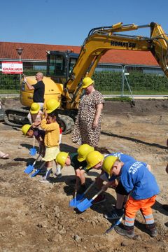 Børn iført hjelme graver i jorden med små skovle foran en gravemaskine.