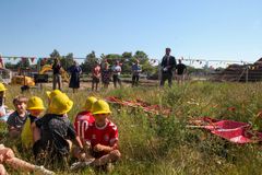 Børn iført gule sikkerhedshjelme sidder på græsset, mens voksne står i baggrunden.