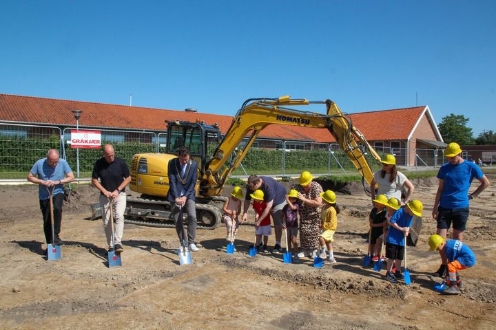 I et byggefelt står en gravemaskine i baggrunden og i forgrunden voksne og børn, der med hver deres spade tager et spadestik.