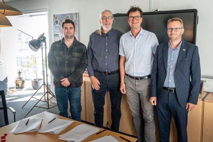 Fire mænd smiler for enden af et bord med underskrevne dokumenter foran sig.