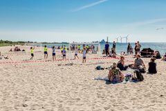 Børn spiller håndbold på en solrig strand, mens folk sidder og nyder solen. Vindmøllerses i baggrunden.
