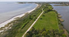 Luftfoto af en smal landtange med veje, græs, sandstrande og vand på begge sider.