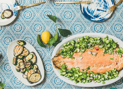 Et bord med en stor stegt laks på et fad, omringet af friske agurker og salat. Ved siden af er der en tallerken med grillede zucchini-skiver og en citron.
