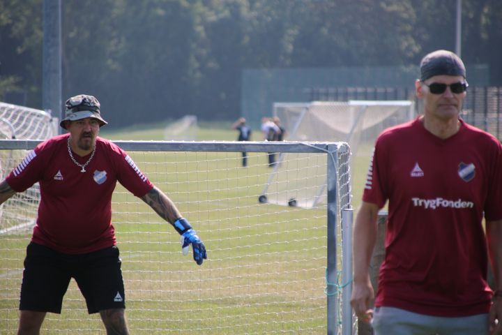 To OMBOLD-spillere i røde trøjer spiller fodbold på en udendørs bane.