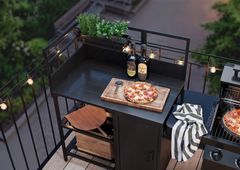 Alt tekst: Udendørs køkkenbord med krydderurter, pizza på skærebræt, to vinflasker og lyskæde.