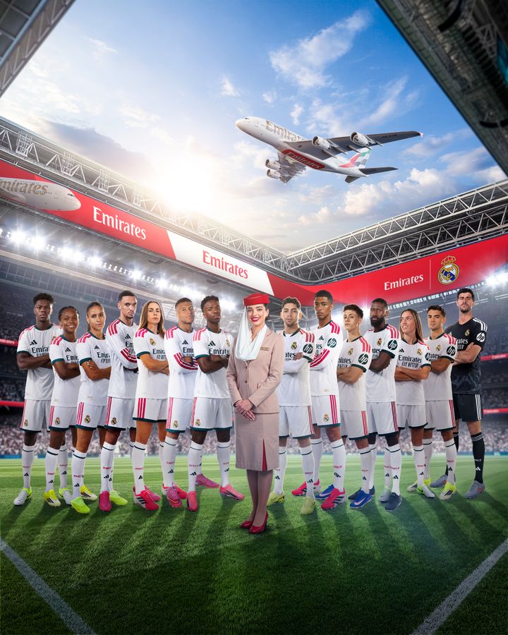 Real Madrids fodboldhold står opstillet på stadion sammen med en kabinemedarbejder fra Emirates, mens et Emirates-fly passerer over stadionet.