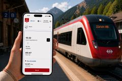 En hånd holder en smartphone med Emirates Skywards Rail-bookingsiden på en togstation i bjergene, mens et moderne rød-hvidt tog holder ved perronen i baggrunden.