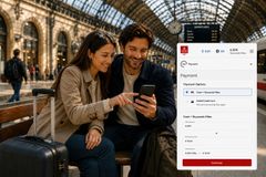 Et par sidder sammen på en bænk på en europæisk togstation og ser på en smartphone med Emirates Skywards Rail-bookingsiden, hvor togrejser kan betales med en kombination af kontanter og Skywards Miles.