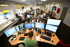 Billede fra kontor på Region Midtjyllands alarmcentral for akut medicinsk hjælp. Hver medarbejder har fem skærme foran sig. Forreste medarbejder sidder med ryggen til fotografen. Hans skærme viser blandt andet et regionskort med veje, en ambulance og lister med informationer, der ikke kan aflæses på billedet. Han har en blok, fire kuglepenne, to tastaturer, to stationære telefoner og en manual liggende på sit skrivebord.