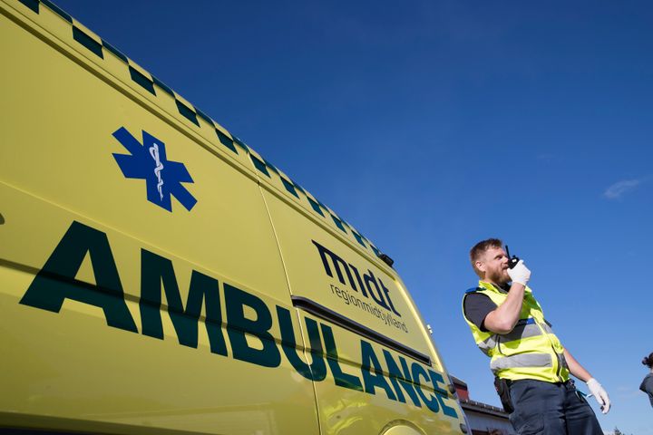 Ambulancebehandler iført gul sikkerhedsvest og latex-handsker står foran ambulance fra Region Midtjylland og taler i radiomodtager.