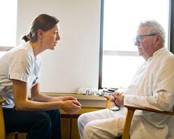 Ældre mand i hospitalstøj sidder i en stol på en hospitalsstue og taler med en behandler i uniform.