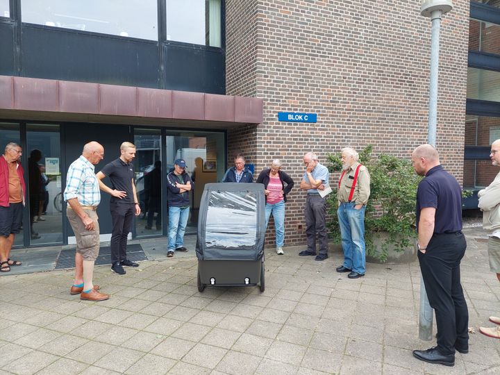 Gruppe mennesker samlet omkring en delecykel foran en bygning.
