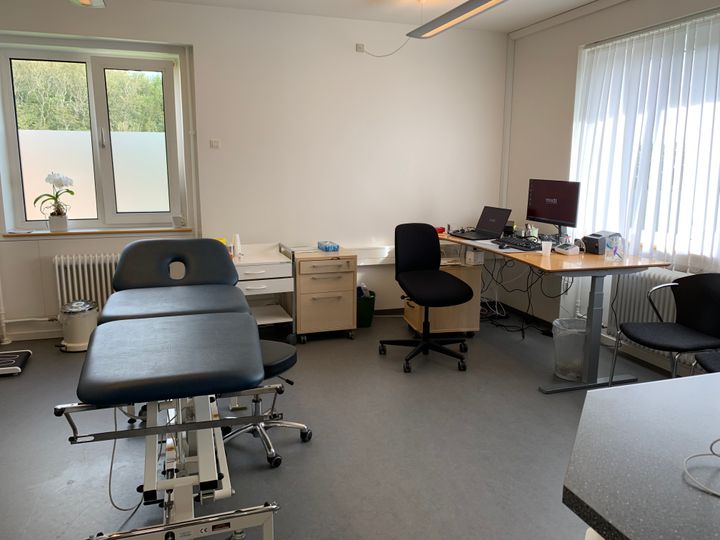 Billedet viser en praktiserende læges lyse undersøgelsesrum, med briks, skrivebord, stol og computer.