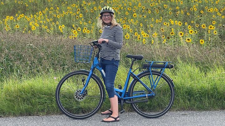 En kvinde med hjelm står med en blå elcykel foran en mark med solsikker.